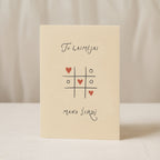 Romantic greeting card featuring a tic-tac-toe game with heart symbols instead of X and O, and the phrase “Tu laimėjai mano širdį” written below.