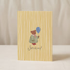 Greeting card featuring a cute teddy bear holding a balloon with the word “SVEIKINU!” on a yellow striped background, cheerful celebration card design.