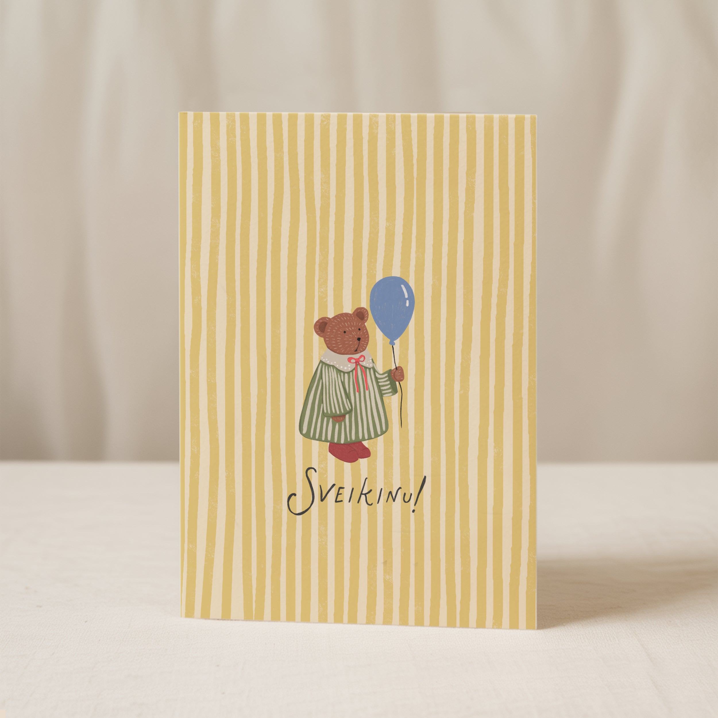 Greeting card featuring a cute teddy bear holding a balloon with the word “SVEIKINU!” on a yellow striped background, cheerful celebration card design.