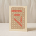 Minimalist greeting card featuring a letter puzzle crossword design with highlighted words “Namai ten kur mama” on a clean white background.