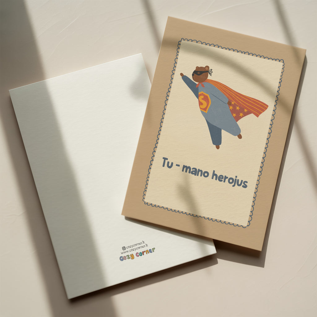 Cute greeting card featuring a teddy bear flying like a superhero with the text “Tu – mano herojus” below, whimsical hero-themed illustration.