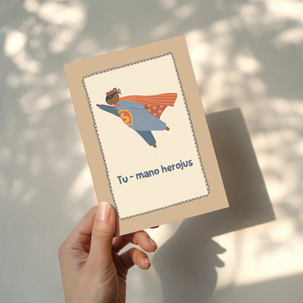 Cute greeting card featuring a teddy bear flying like a superhero with the text “Tu – mano herojus” below, whimsical hero-themed illustration.