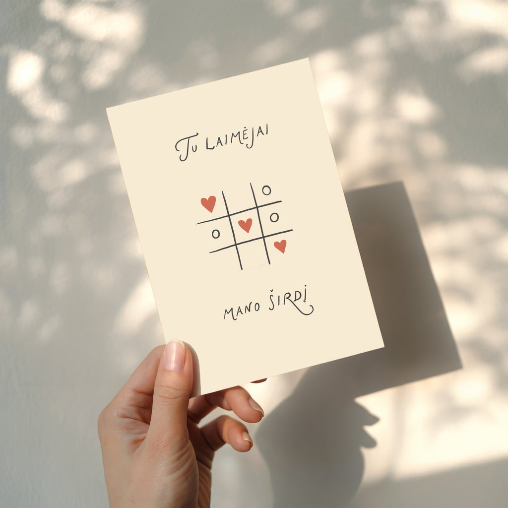 Romantic greeting card featuring a tic-tac-toe game with heart symbols instead of X and O, and the phrase “Tu laimėjai mano širdį” written below.