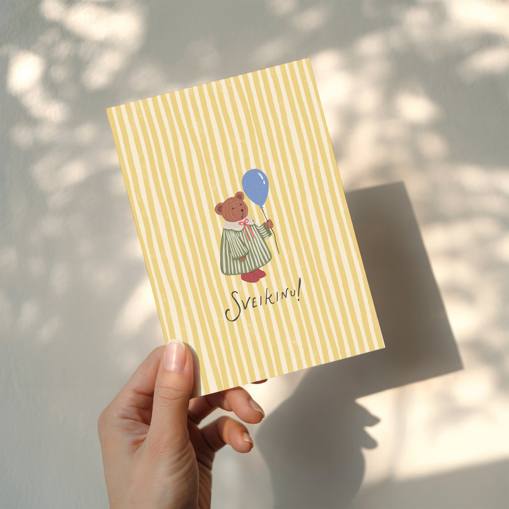 Greeting card featuring a cute teddy bear holding a balloon with the word “SVEIKINU!” on a yellow striped background, cheerful celebration card design.