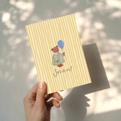 Greeting card featuring a cute teddy bear holding a balloon with the word “SVEIKINU!” on a yellow striped background, cheerful celebration card design.