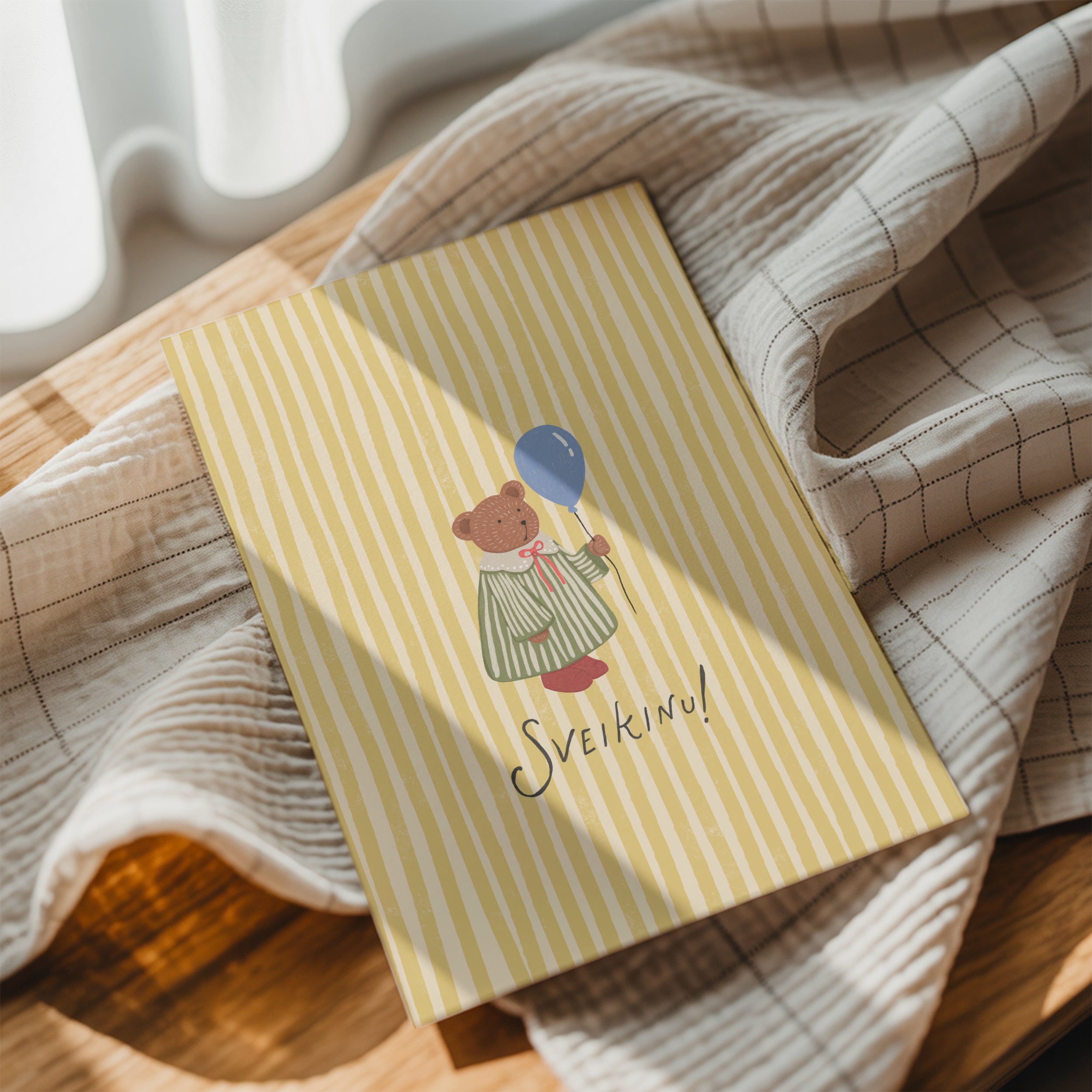 Greeting card featuring a cute teddy bear holding a balloon with the word “SVEIKINU!” on a yellow striped background, cheerful celebration card design.